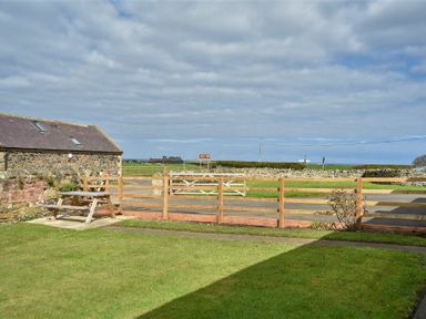 An outdoor area with a wooden picnic table a stone building and wooden fence with open fields beyond at Dryden Cottage in Bamburgh