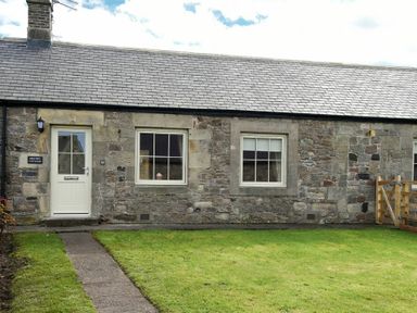 The front exterior of a stone cottage with two windows a door and a grass lawn at Dryden Cottage in Bamburgh