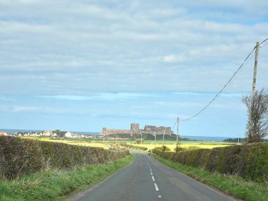 A rural road between hedges leading to a castle with fields and houses at Dryden Cottage in Bamburgh