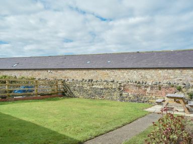 A grassy backyard with a wooden picnic table near a stone wall and a long stone building at Dryden Cottage in Bamburgh