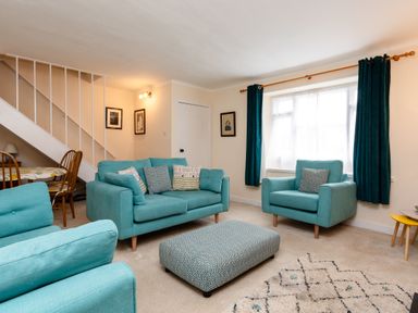 A living room with turquoise sofas a patterned ottoman curtains and a dining table under stairs at Percy Cottages No4 in Alnmouth