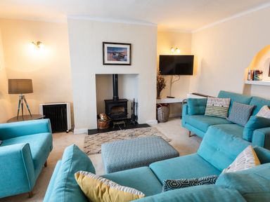 A living room with turquoise sofas a patterned ottoman a wood-burning stove and a wall-mounted TV at Percy Cottages No4 in Alnmouth