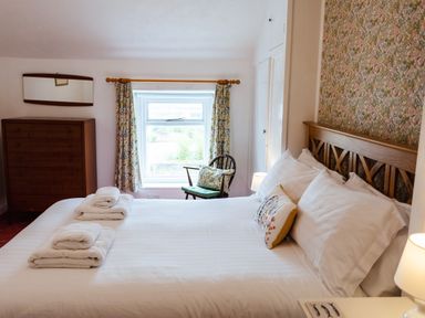 A bedroom with a double bed with white linens folded towels on the bed a wooden chest of drawers a floral curtain and a chair by the window at Percy Cottages No4 in Alnmouth