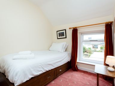 A bedroom with a single bed with white bedding a wooden side table with a lamp and a window with red curtains at Percy Cottages No4 in Alnmouth