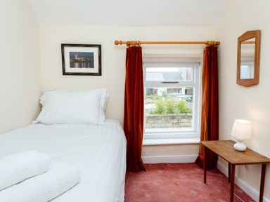 A small bedroom with a single bed white bedding towels on bed orange curtains and a wooden table with lamp at Percy Cottages No4 in Alnmouth