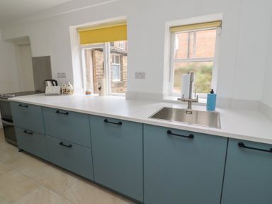 A kitchen with blue cabinets white countertops a stainless steel sink and two windows with yellow blinds at Percy Cottages No4 in Alnmouth