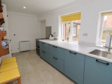 A kitchen with blue lower cabinets white countertops yellow window blinds stove and wooden chairs with yellow cushions at Percy Cottages No4 in Alnmouth