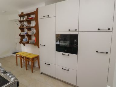 A kitchen with white cabinets a built in black microwave wooden shelves with dishes and three wooden stools with yellow cushions at Percy Cottages No4 in Alnmouth