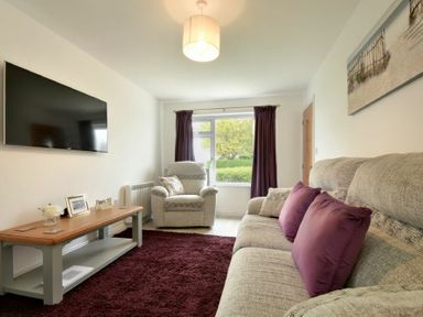 A living room with a beige sofa with purple cushions a beige armchair a wooden coffee table and a wall mounted television at The Chevy in Seahouses