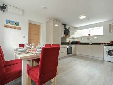 A kitchen with white cabinets, a washing machine, dining table with red chairs, and a wooden door at The Chevy in Seahouses