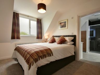A bedroom with a bed and plaid cushions and blanket next to a window and a door to a bathroom at The Chevy in Seahouses