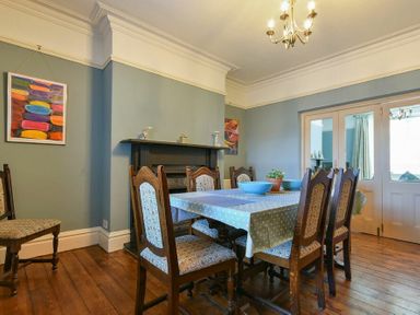 A dining room with a wooden table and six upholstered chairs a fireplace and paintings on blue walls at Nettie's View in Amble