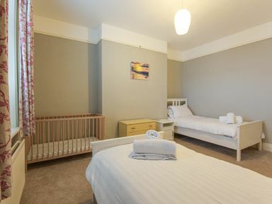 A bedroom with two single beds a wooden crib a chest of drawers and a window with floral curtains at Nettie's View in Amble