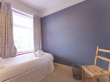 A bedroom with a single bed white linens rolled towels floral curtains a wooden chair and a trash bin at Nettie's View in Amble