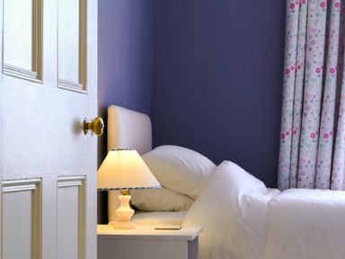 A bedroom with a white door partially open a bed with white bedding a bedside table with a lamp and patterned curtains at Nettie's View in Amble