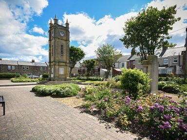 A clock tower in a garden with flowers trees and brick houses in the background at Netties View in Amble