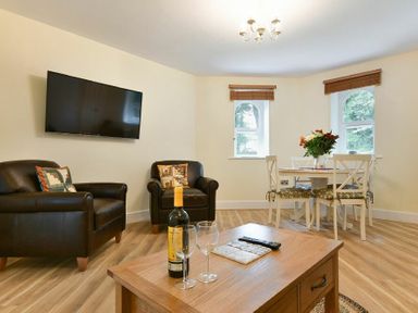 A living room with two black leather armchairs a mounted flat screen TV a wooden coffee table and a dining table with four chairs and flowers at The Lodge (Alnwick) in Alnwick