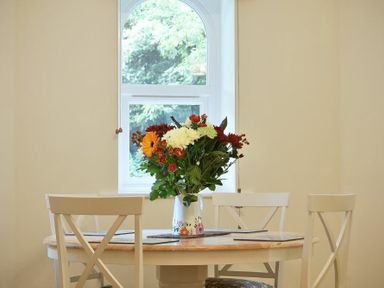 A round wooden dining table with a vase of flowers and three white chairs near a window at The Lodge (Alnwick) in Alnwick