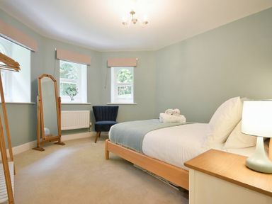 A bedroom with a bed side table lamp chair full length mirror and three windows at The Lodge (Alnwick) in Alnwick