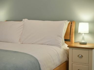 A bed with white pillows and blanket next to a wooden nightstand with a lamp at The Lodge (Alnwick) in Alnwick