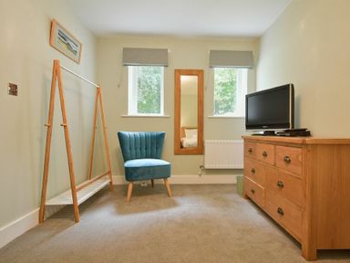A room with a blue chair a wooden dresser with a TV a wooden clothes rack and two windows at The Lodge (Alnwick) in Alnwick