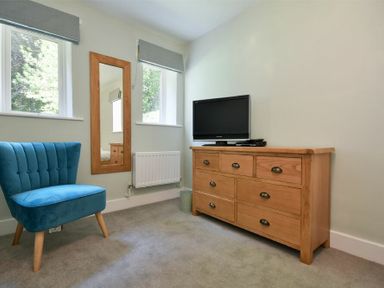 A room with a blue chair wooden dresser with a television on top and two windows at The Lodge (Alnwick) in Alnwick