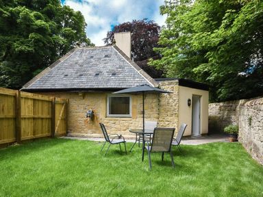 A backyard with green grass a round table with four chairs and an umbrella beside a house with stone walls and a sloped roof at The Lodge (Alnwick) in Alnwick