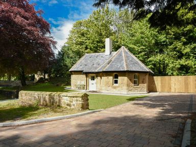 A small stone cottage with a pitched roof surrounded by trees and a paved driveway at The Lodge (Alnwick) in Alnwick