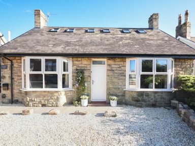 The front exterior of a stone cottage with two bay windows and a white door at Marmalade Cottage in Seahouses