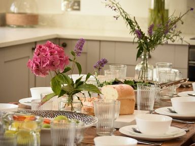 A dining table set with glasses bowls plates a loaf of bread and flowers in a kitchen at Marmalade Cottage in Seahouses