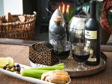 A table with two glasses of red wine a wine bottle a metal container bread celery olives lemon slices and a basket with firewood near a fireplace at Marmalade Cottage in Seahouses