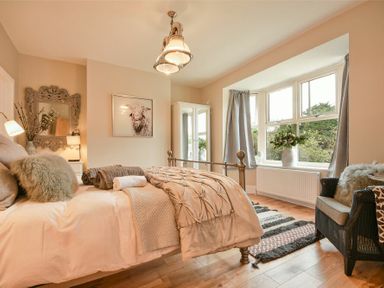 A bedroom with a metal bed frame, a large window with curtains, a chair, a mirror, and a wardrobe at Marmalade Cottage in Seahouses