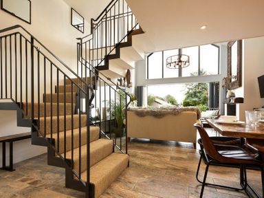 A living area with a staircase carpeted in beige a beige sofa a wooden dining table with chairs and large windows showing greenery at Marmalade Cottage in Seahouses