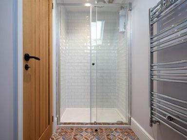 A shower enclosure with glass doors and white tiled walls next to a wooden door and a metal towel radiator at Marmalade Cottage in Seahouses