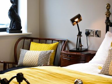 A bedroom corner with a wooden chair with yellow and patterned cushions a bedside table with a lamp and part of a bed with yellow bedding at Marmalade Cottage in Seahouses
