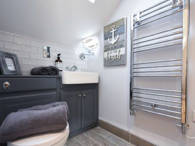 A bathroom with a sink, gray cabinets, folded towels, a wall mirror, a towel radiator, and a beach themed wall sign at Marmalade Cottage in Seahouses