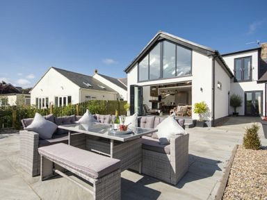 An outdoor patio area with a wicker corner sofa and glass table in front of a house with large windows at Marmalade Cottage in Seahouses