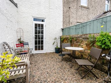 A small outdoor patio with a wooden bench table and chairs outside a white building at Anchor Cottage Seahouses