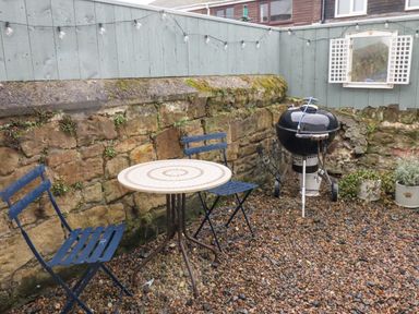 An outdoor patio with two blue metal chairs a round table a black barbecue grill gravel ground stone wall and wooden fence at Anchor Cottage (Seahouses) in Seahouses