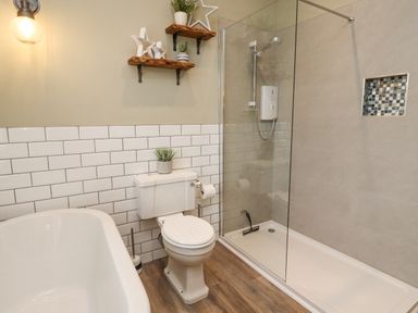 A bathroom with a white bathtub a toilet a walk in shower and wooden shelves on the wall at Anchor Cottage Seahouses