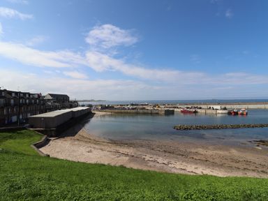 A small harbor with boats docked and a sandy beach with green grass in the foreground at Anchor Cottage in Seahouses