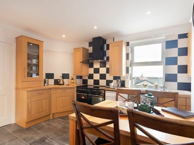 A kitchen with wooden cabinets a black and white checkered backsplash a black stove a wooden dining table with chairs and a window at Church View in Bamburgh