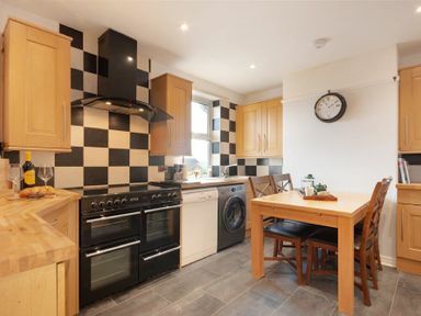 A kitchen with wooden cabinets, a black stove, checkered black and white backsplash, a wooden dining table with chairs and a clock on the wall at Church View in Bamburgh