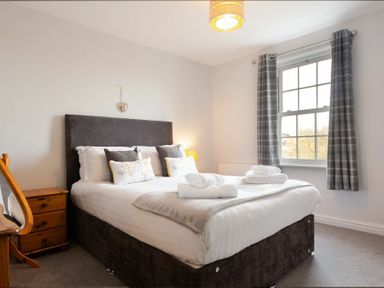 A bedroom with a double bed bedside table armchair and window with checkered curtains at Church View in Bamburgh