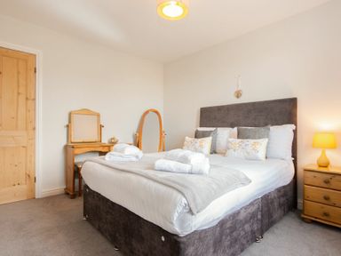 A bedroom with a double bed pillows and towels a dressing table with a mirror a standing mirror a bedside table with a lamp and a wooden door at Church View in Bamburgh