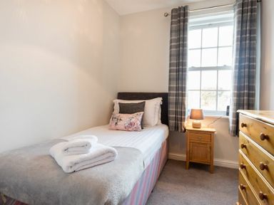A bedroom with a single bed side table with a lamp window with curtains and a chest of drawers at Church View in Bamburgh