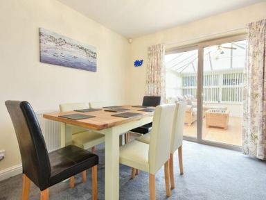 A dining room with wooden table and mixed color chairs next to sliding glass doors and floral curtains at Annstead House in Seahouses