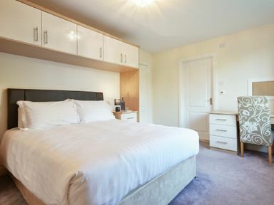 A bedroom with a double bed white bedding overhead cabinets a bedside table and a desk with a patterned chair at Annstead House in Seahouses