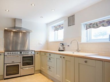 A kitchen with a stainless steel stove and hood a kettle on the countertop and twin windows with floral blinds at Cargoes in Craster