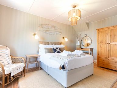 A bedroom with a double bed, wicker chair, wooden wardrobe, small wooden nightstand, and a desk with a round mirror at Cargoes in Craster